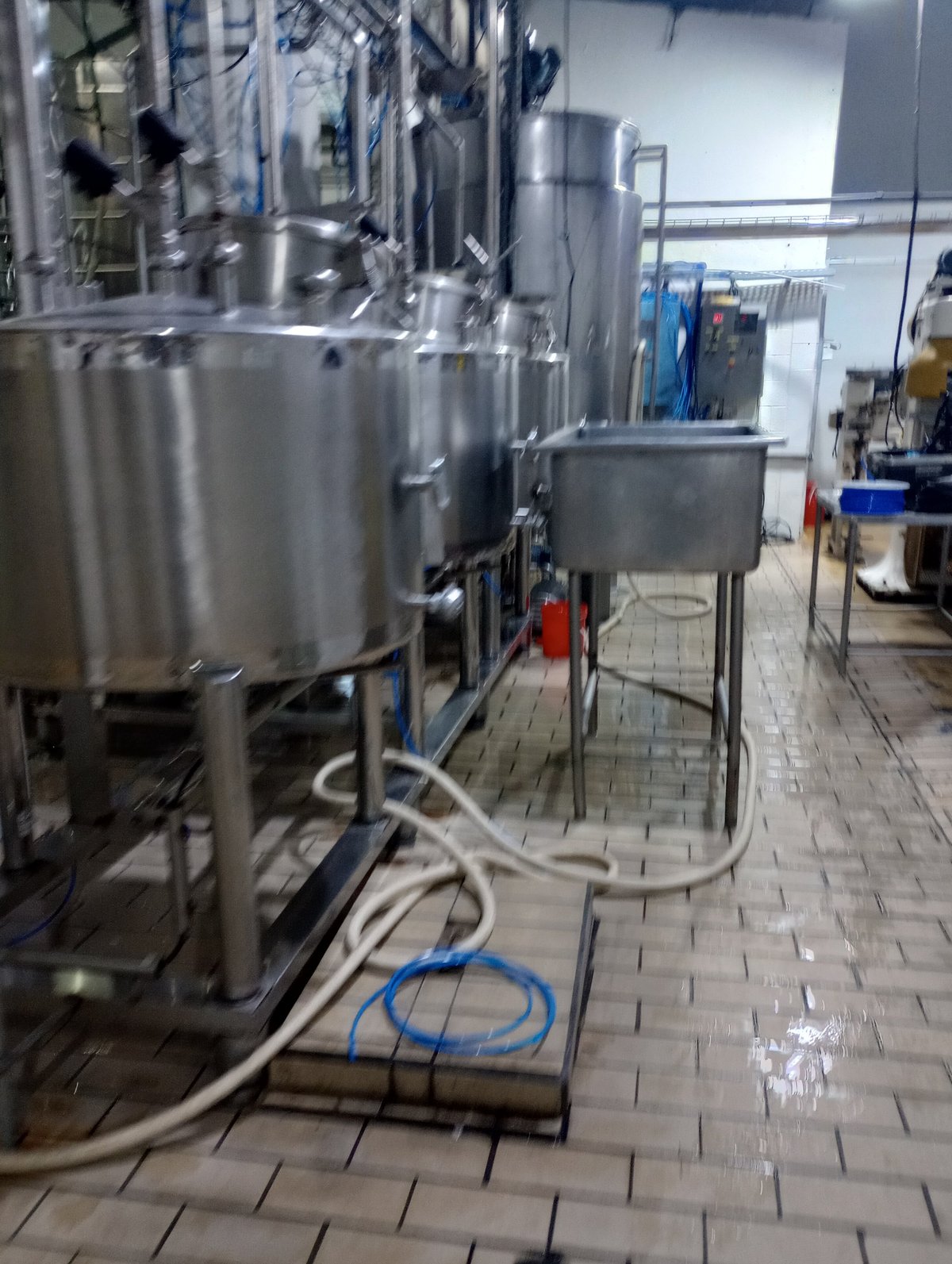 Industrial stainless steel tanks and equipment in a facility with concrete flooring and blue hoses connected to machinery