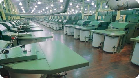 Industrial textile factory floor with rows of large green machinery and work tables under bright overhead lighting